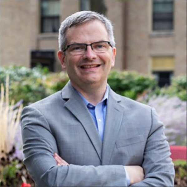 A middle-aged man with short gray hair and glasses, wearing a light gray suit and blue shirt.