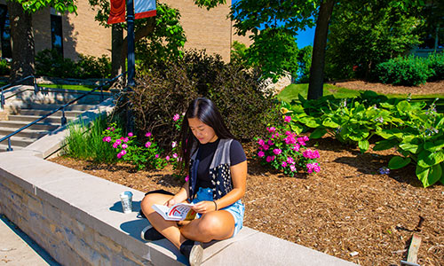 a Carroll University student sitting on a ledge reading a book.