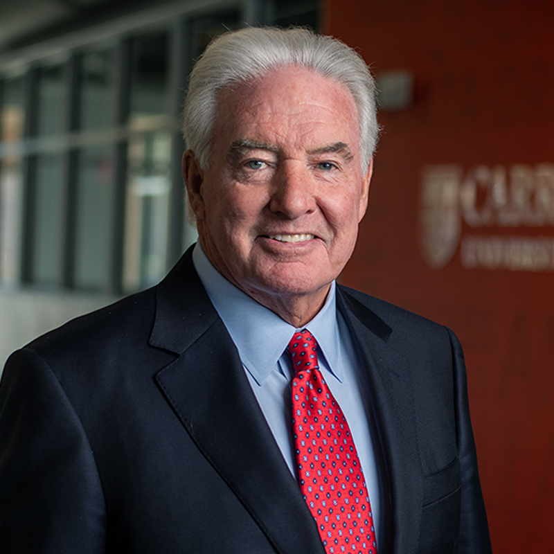 Older man with white hair in a suit and red tie, smiling in front of a Carroll University backdrop.