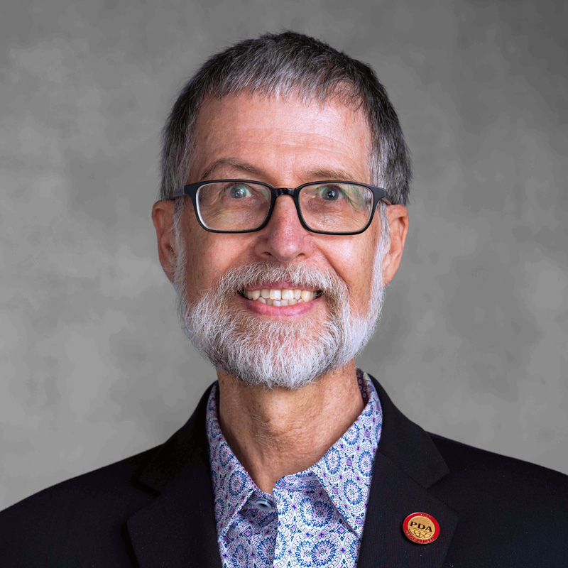 A smiling older man with glasses, a gray beard, and short hair wears a patterned shirt and black blazer with a red "PDA" pin on the lapel.
