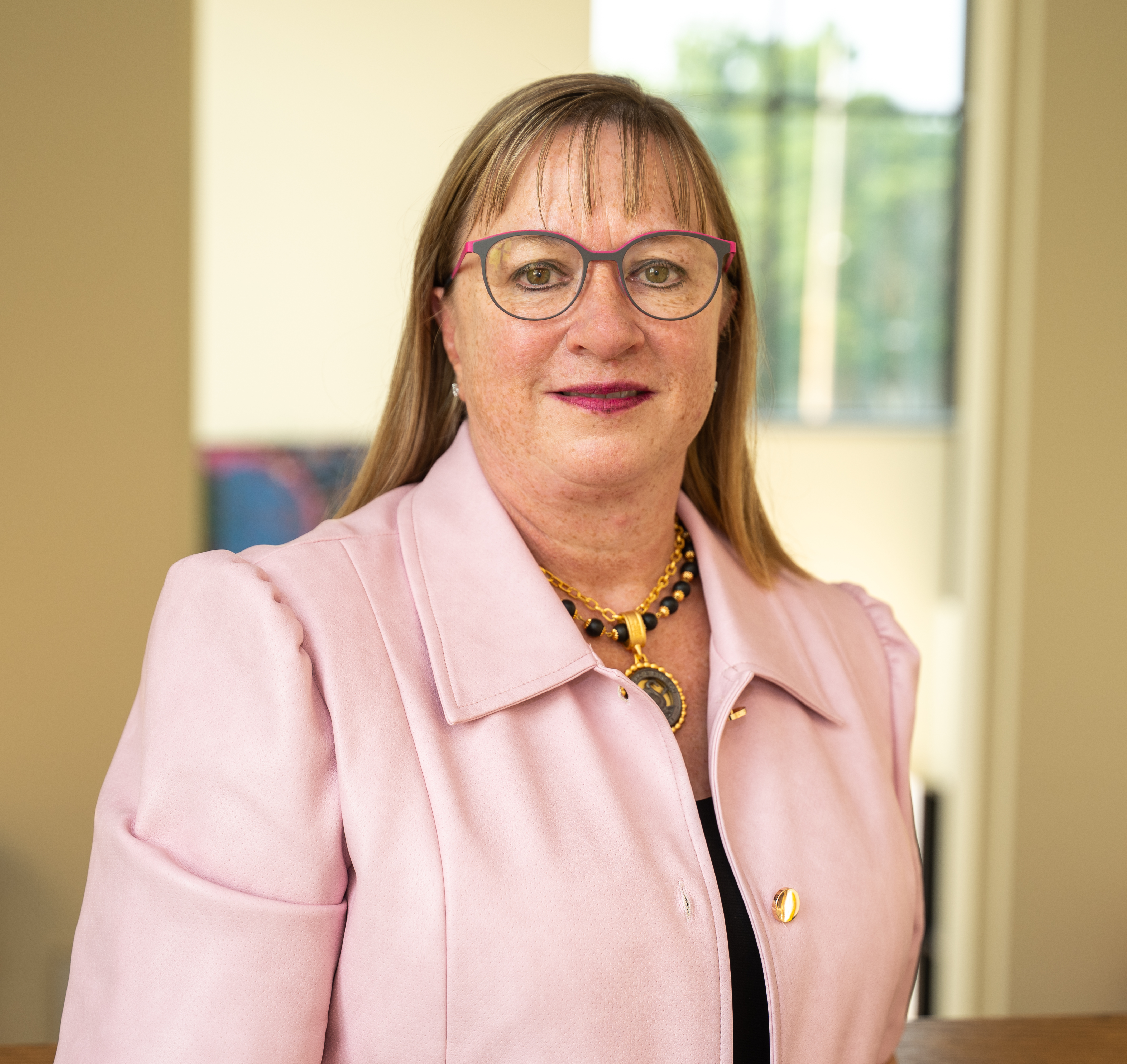 A person in a light pink jacket and necklace stands indoors near a window.