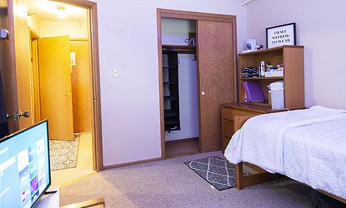 A tidy, modest bedroom with an open closet, a well-organized dresser, and a bed, with an open door leading to the hallway, and a television screen displaying a menu interface in the foreground.