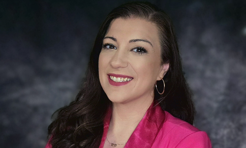 Professional portrait photograph of a woman with long dark brown hair wearing a vibrant pink blazer over a white top. 