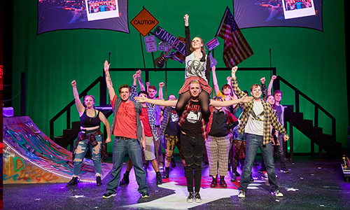 A large ensemble of performers on a brightly lit stage raise their fists in a high-energy moment, with protest-style signs and an American flag visible against a green backdrop.