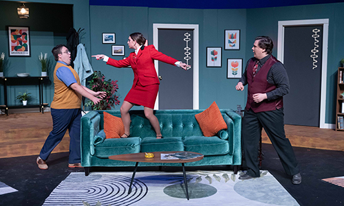 A woman in a red suit stands on a couch pointing assertively at a man while another man looks on in a staged living room scene.