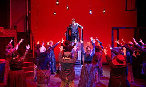 A stage scene shows a central performer elevated above a crowd of actors reaching upward under dramatic red lighting.