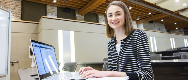a Carroll University student smiling at the camera and working on a laptop computer.