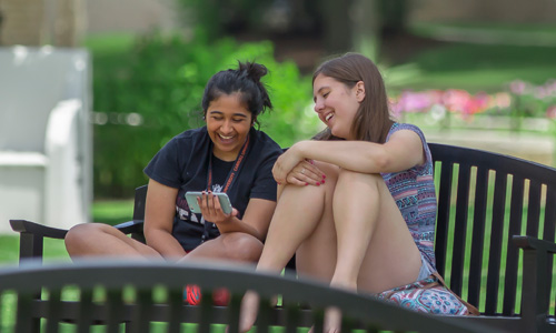two Carroll students sitting on a bench laughing and looking at a cell phone.