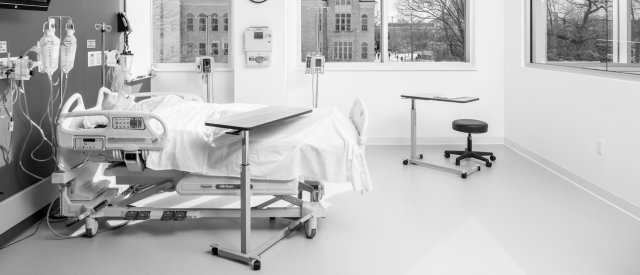 A monochrome photograph of a modern, empty hospital room with a bed, various medical equipment, and a large window overlooking a building outside.