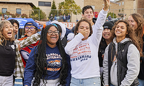 A group of Carroll University students laugh and cheer together outdoors during Homecoming, wearing Carroll apparel and fall clothing.