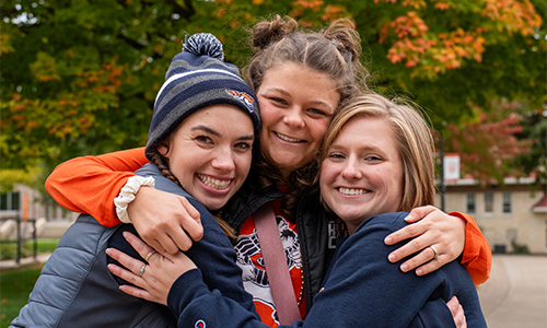 Three young women in matching navy and orange jackets embrace and smile at the camera outdoors on an autumn day, with colorful fall foliage visible in the background. 