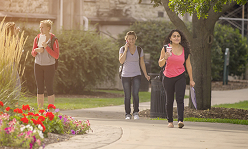 Carroll University students walking along a campus pathway on a sunny day, with flowers and trees in the background.